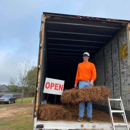 Richard in an orange shirt loading bales out of a trailer next to an OPEN sign